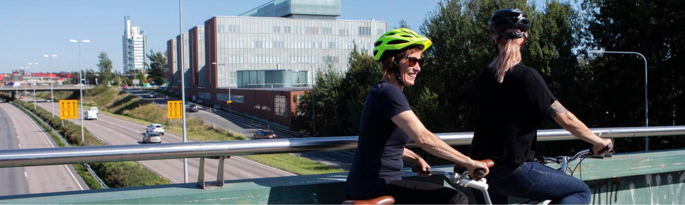 Kaksi henkilöä kypärät päässä tandemin päällä pysähtyneinä sillalle, taustalla suuri autotie ja Iiris-keskus.